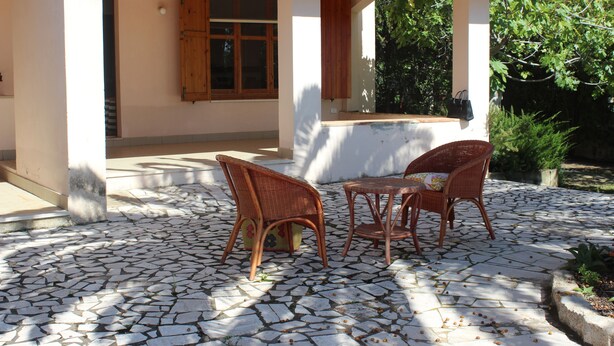 Un espace extérieur abrité, aménagé avec deux fauteuils en osier et une petite table, entouré d’une terrasse carrelée en pierre.