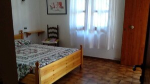 Une chambre à coucher avec un lit en bois, une chaise, une petite table, une armoire et une fenêtre avec des rideaux.