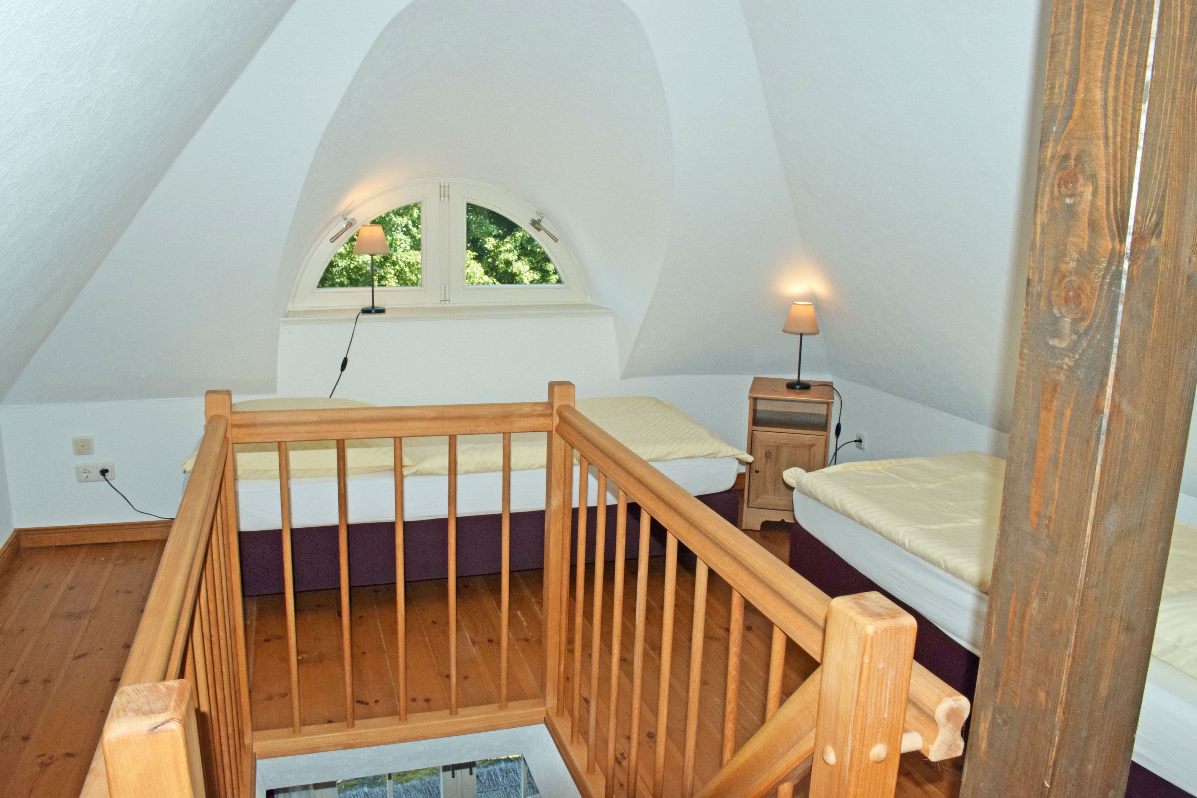 A bedroom with a wooden staircase, a window with a view of trees, a bedside table with a lamp, and a bed with a headboard.