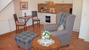 A cozy living room with a grey armchair, a plaid ottoman, and a wooden coffee table with a floral centerpiece. A dining table with chairs is positioned in the background, and a kitchen area is visible with white cabinets and a wooden backsplash. - 07 Vacation apartment - Karolas Landhus under the thatched roof (Mönchgut)
