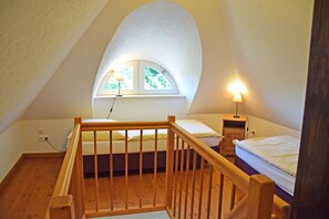 A small, cozy attic bedroom with a sloped ceiling, a wooden staircase, two single beds, a small window with a view of trees, and a bedside table with a lamp.
