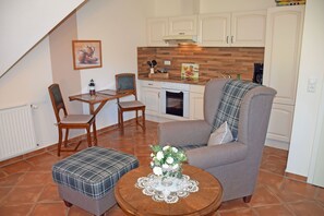A cozy living room with a grey armchair, a plaid ottoman, and a wooden coffee table with a floral centerpiece. A dining table with chairs is positioned in the background, and a kitchen area is visible with white cabinets and a wooden backsplash. - 07 Vacation apartment - Karolas Landhus under the thatched roof (Mönchgut)