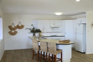 A kitchen with white cabinets, a central island with bar stools, a sink, and a refrigerator. - Beaches 1 - 1/7 Convent (Yamba)