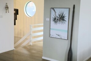 A hallway with a staircase, a framed artwork of a tree and beach, and a tall wooden sculpture. - Beaches 1 - 1/7 Convent (Yamba)