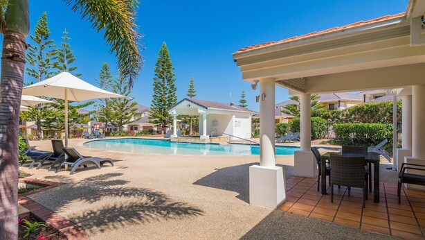 A pool area with lounge chairs, a covered seating area, and a view of residential buildings in the background. - Breakers 2/2 (Yamba)