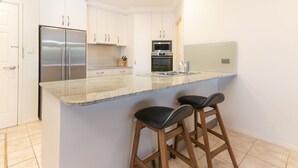 A modern kitchen with a stainless steel refrigerator, double ovens, a microwave, and a granite countertop. There are two wooden bar stools at the island. - Breakers 2/2 (Yamba)