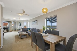 A spacious living area with a dining table, chairs, sofas, ceiling fans, and large windows offering a view. - Breakers 2/12 (Yamba)