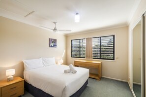 A hotel room with a bed, bedside table, lamp, ceiling fan, window with blinds, and a wooden desk. - Breather 2/44 Beachside Way (Yamba)