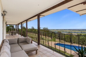 A covered balcony with a sofa, a dining table, and a view of a swimming pool and landscape.