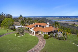 A large, single-story house with a red-tiled roof, surrounded by lush greenery and palm trees, overlooking a coastal landscape.