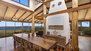 Een ruime eetruimte met een grote houten tafel en stoelen, een hoog plafond met zichtbare balken, en een groot raam met uitzicht op het landschap.