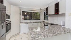 A modern kitchen with white cabinetry, a granite countertop, stainless steel appliances, and a window providing a view of greenery outside. - Oceania Beach Paradise (Yamba)