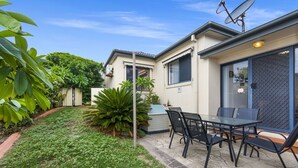 A house with a patio area featuring a table and chairs, surrounded by a garden with various plants. - Oceania Beach Paradise (Yamba)