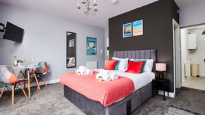A modern hotel room with a large bed, a TV, a dining area with chairs, and a view of Prague. - Stylish Apt Near EMA; Gym & Pool Access, Fast WiFi (Castle Donington)