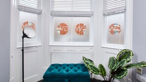 A room with a teal tufted bench, a floor lamp, and a potted plant with variegated leaves. - Stylish Apt Near EMA; Gym & Pool Access, Fast WiFi (Castle Donington)