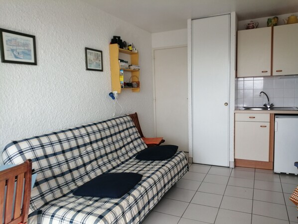 A small, furnished room with a sofa, a chair, a kitchen area with cabinets and a sink, and a door leading to another room.