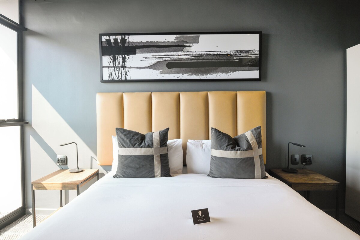 Ein modernes Hotelzimmer mit einem großen Bett, einem Kopfteil, zwei Nachttischen, einer Nachttischlampe, einem gerahmten abstrakten Gemälde und einem kleinen Schild auf dem Bett.