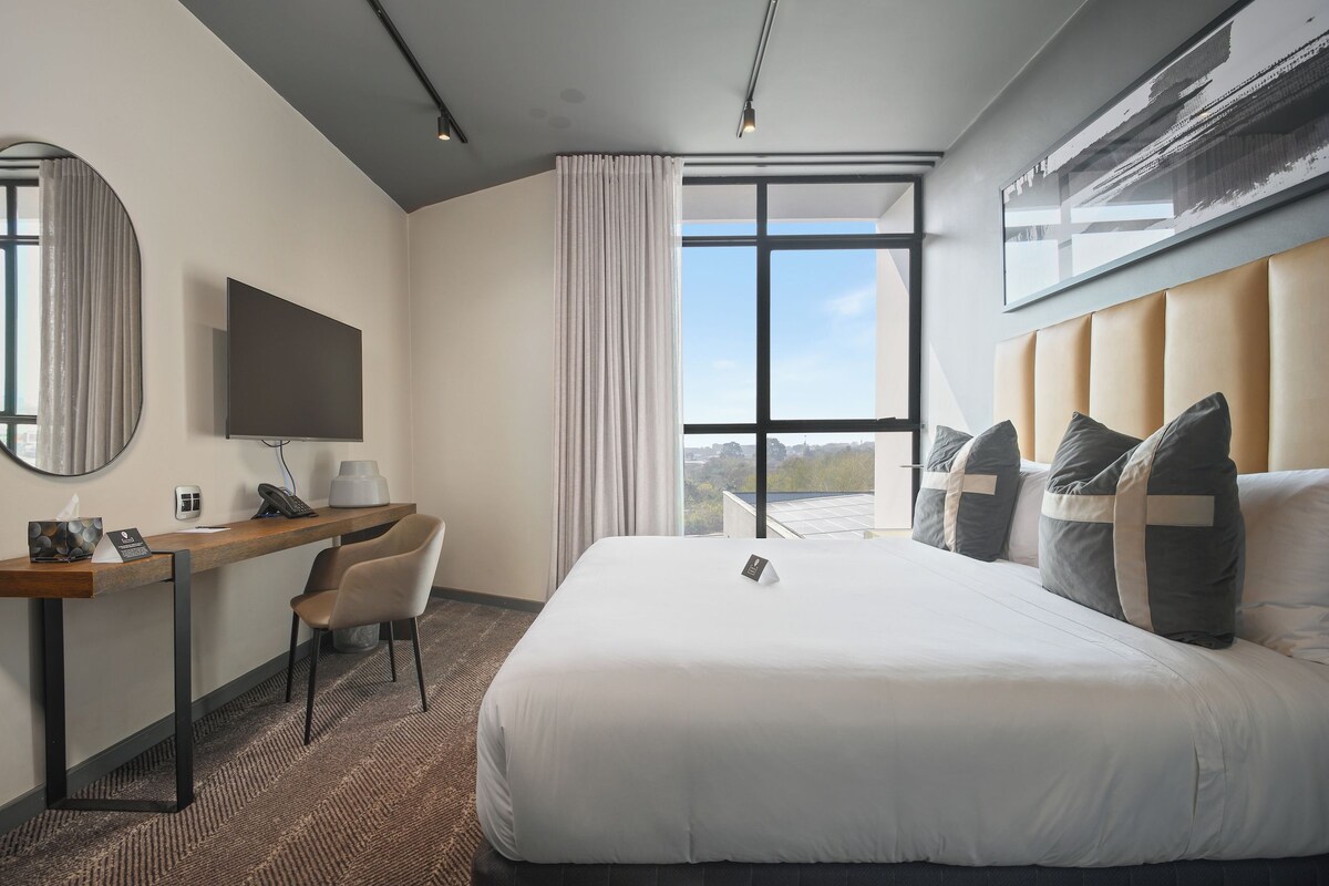 A hotel room with a large bed, a desk with a chair, a flat-screen TV, a large window with curtains, and a view of the city.