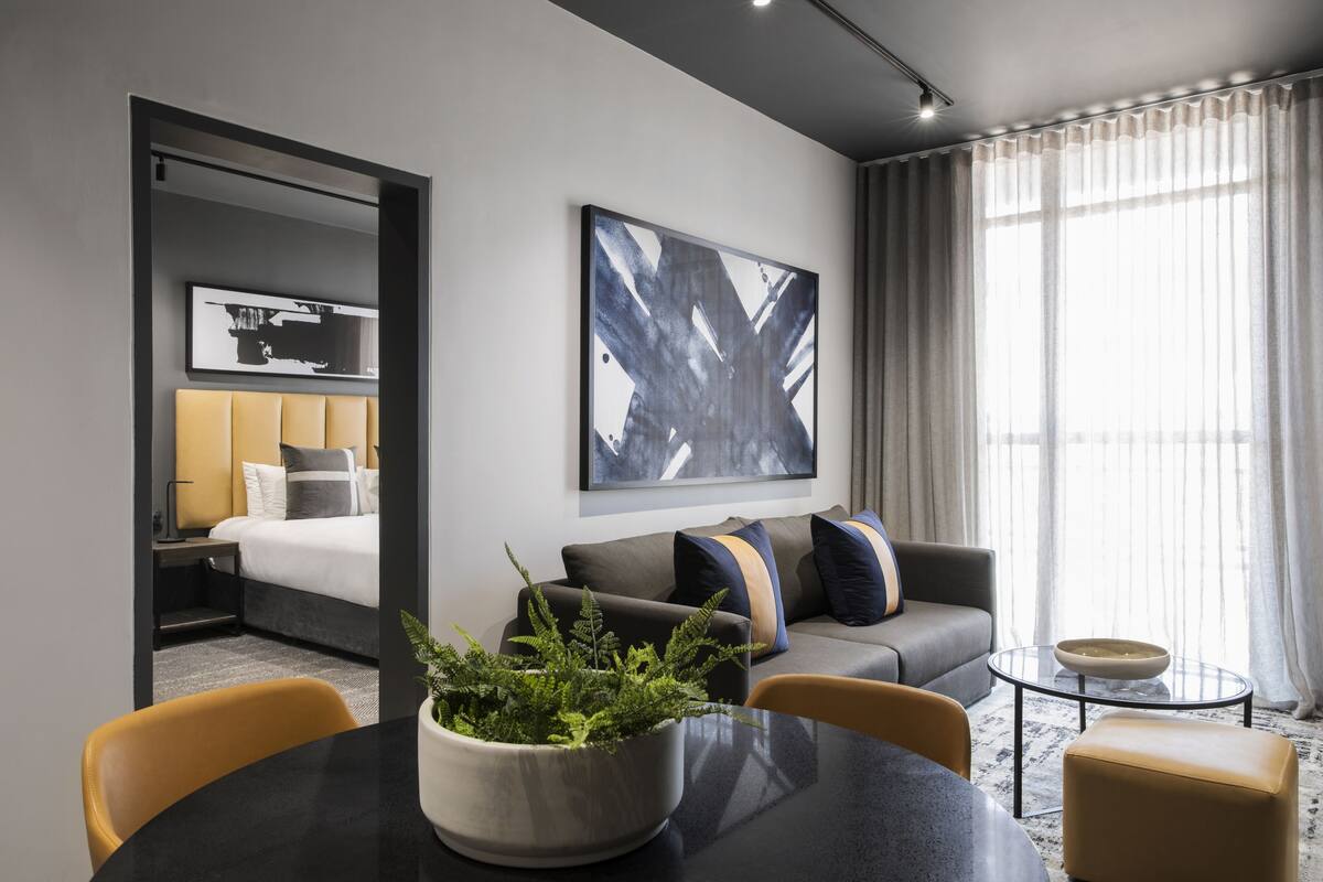 A modern hotel room with a bed, sofa, dining table, and a large window with curtains.