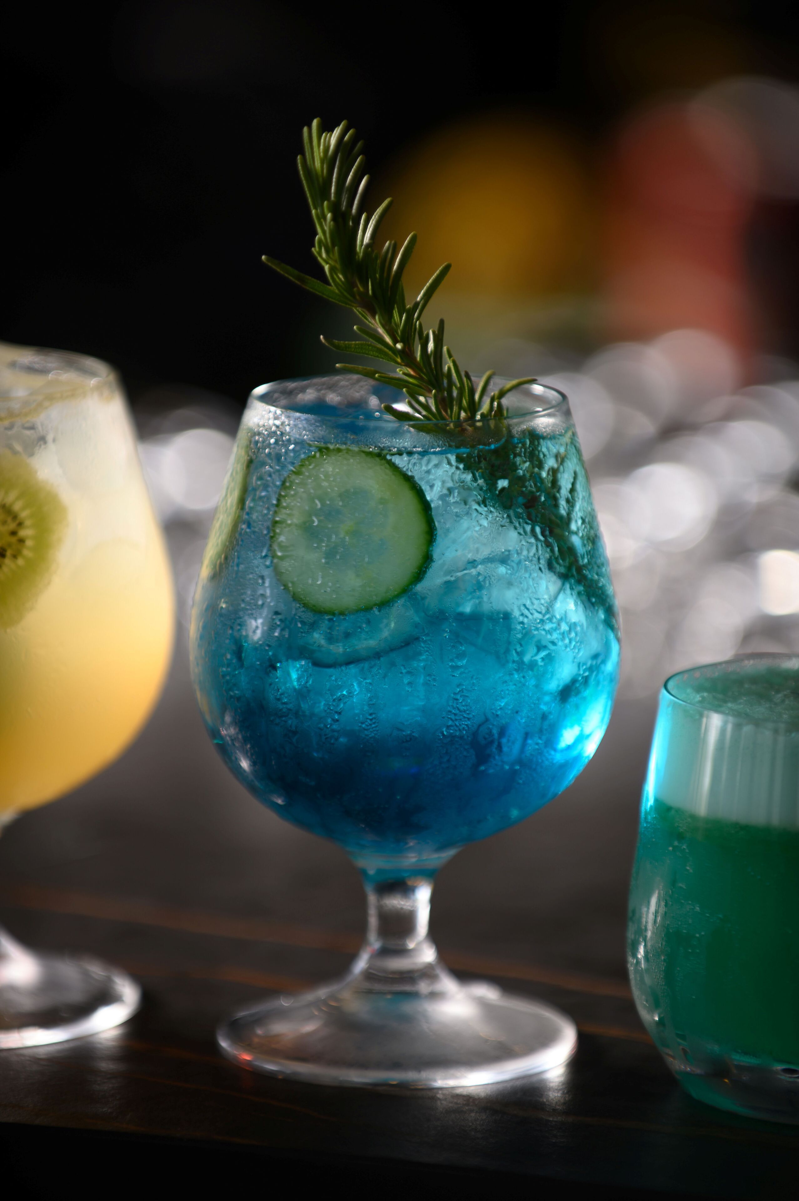 Et cocktailglas med en blå drink, pyntet med rosmarin og et skiver af lime, på en træoverflade.