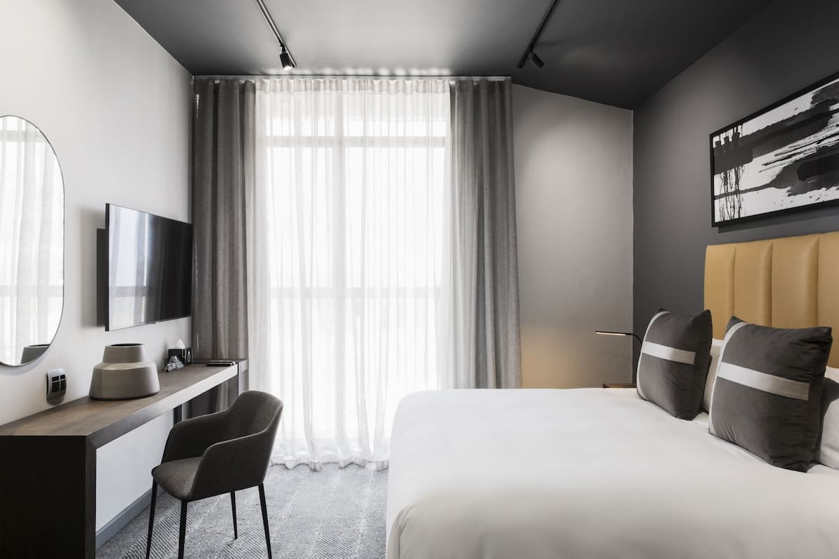 A modern hotel room with a bed, a desk, a chair, a television, and a large window with curtains.