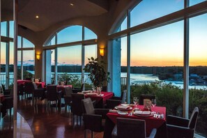Un restaurante con mesas dispuestas para comer, amplias ventanas que ofrecen vista a un cuerpo de agua y un paisaje lejano.