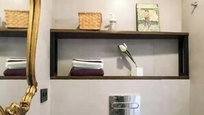 A modern bathroom with a white sink, a toilet, and a shower area. There is a shelf with toiletries and a mirror with a gold frame. - ROYAL OPERA MADRID (Madrid)