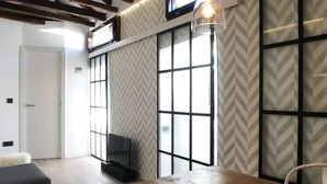 A room with a wooden table and bench, a TV on a stand, and a large window with a chevron-patterned wall. - ROYAL OPERA MADRID (Madrid)