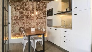 A compact kitchen with white cabinets, a wooden dining table, and a brick wall backdrop. - ROYAL OPERA MADRID (Madrid)