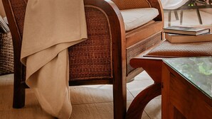 Uma cadeira de vime com almofada e manta, uma luminária de chão e uma mesa de vidro com um livro.