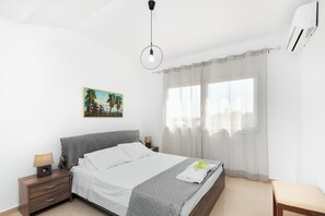 Ein Schlafzimmer mit einem Bett, Nachttischen, einer Pendelleuchte, einem Fenster mit Vorhängen und einer Wand-Klimaanlage.