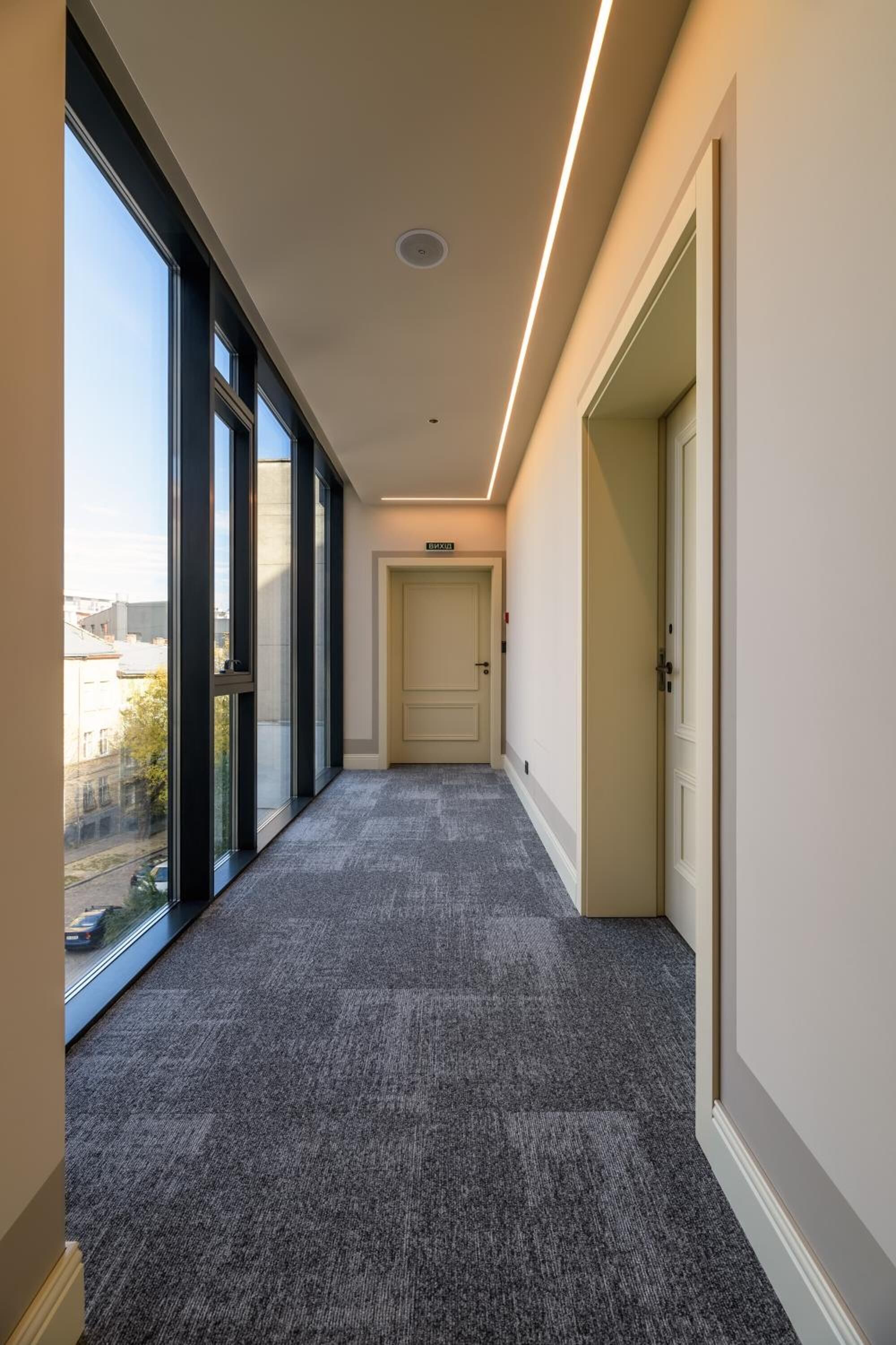 A corridor with large windows, carpeted floor, and a door at the end.