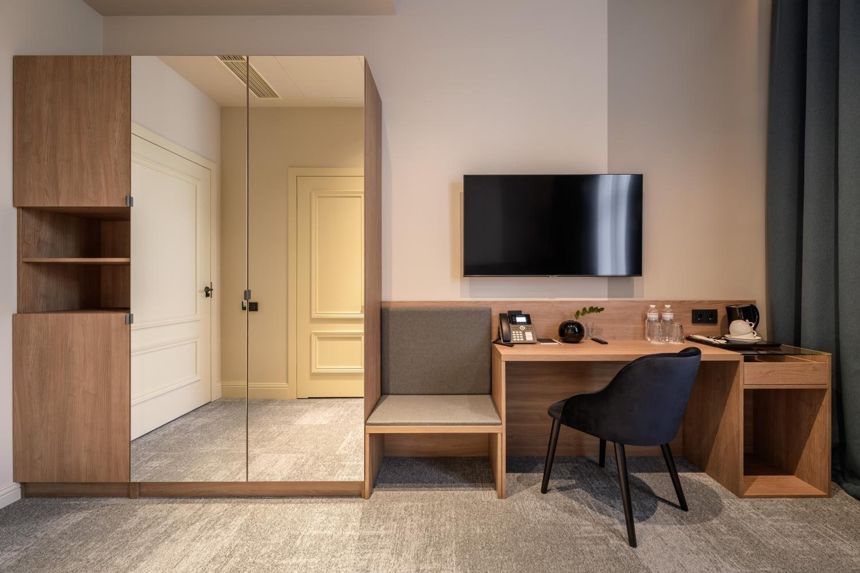 A modern hotel room with a wooden desk, a comfortable chair, a flat-screen TV, and a wardrobe with a mirror.
