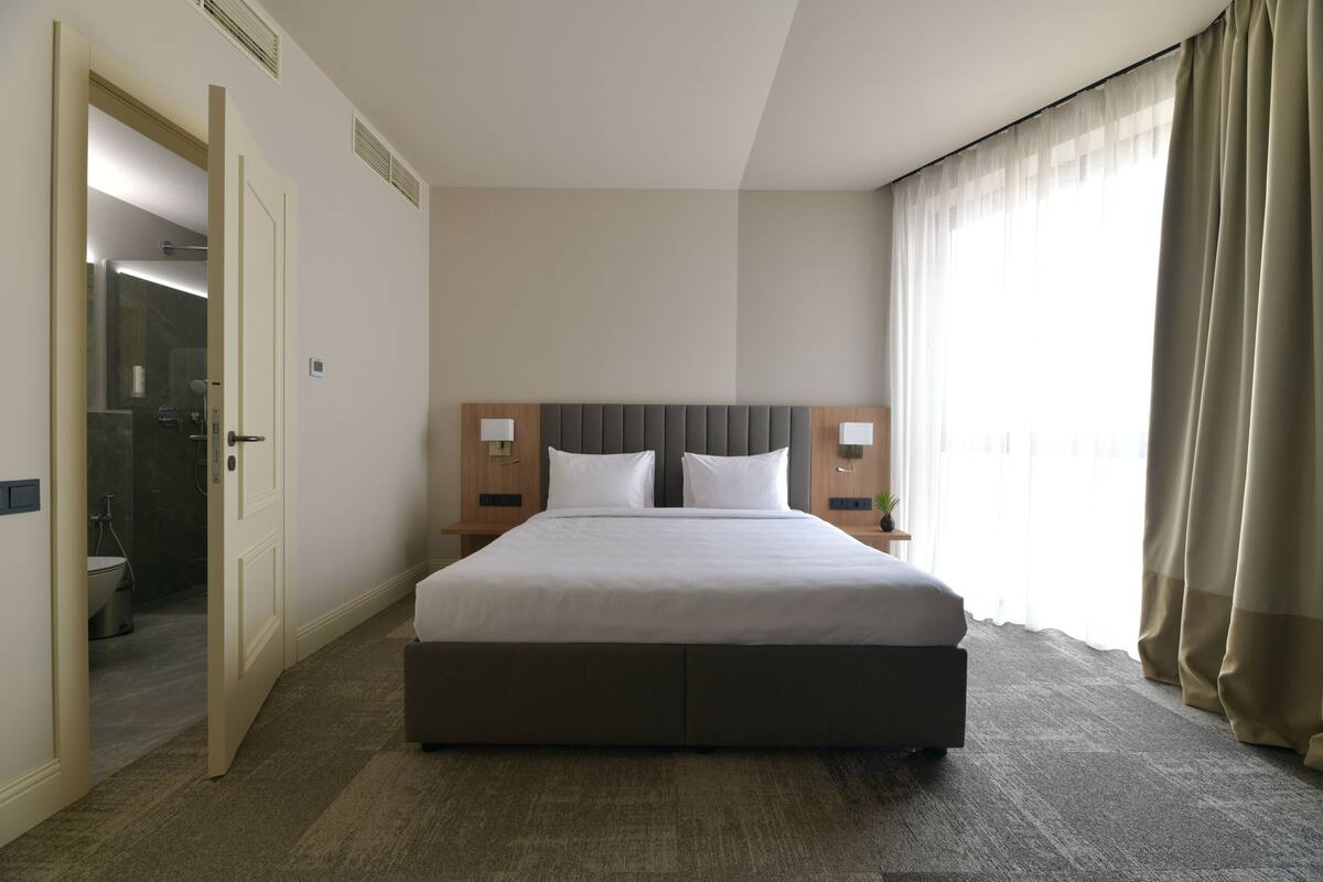 A hotel room with a large bed, a nightstand, a bedside table, a window with curtains, and a door leading to a bathroom.