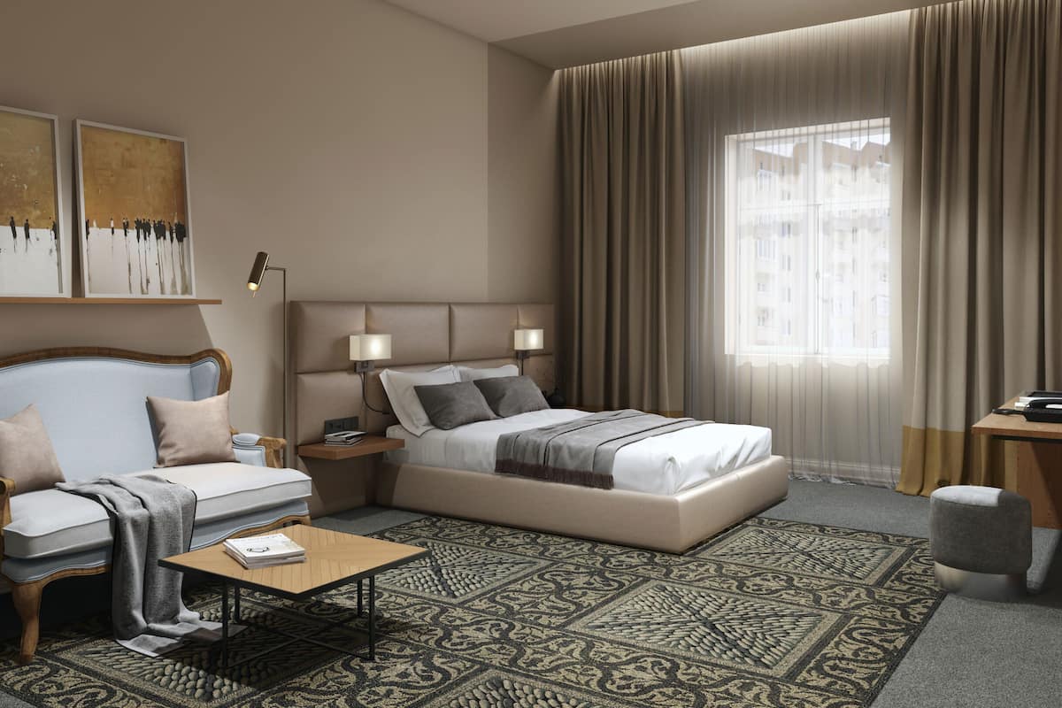 A modern hotel room with a bed, a sofa, a coffee table, and a desk. There are two paintings on the wall, a potted plant, and a window with curtains.