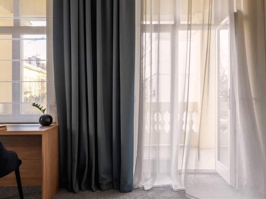 Una habitación con escritorio, silla, ventana grande con cortinas grises y piso alfombrado.