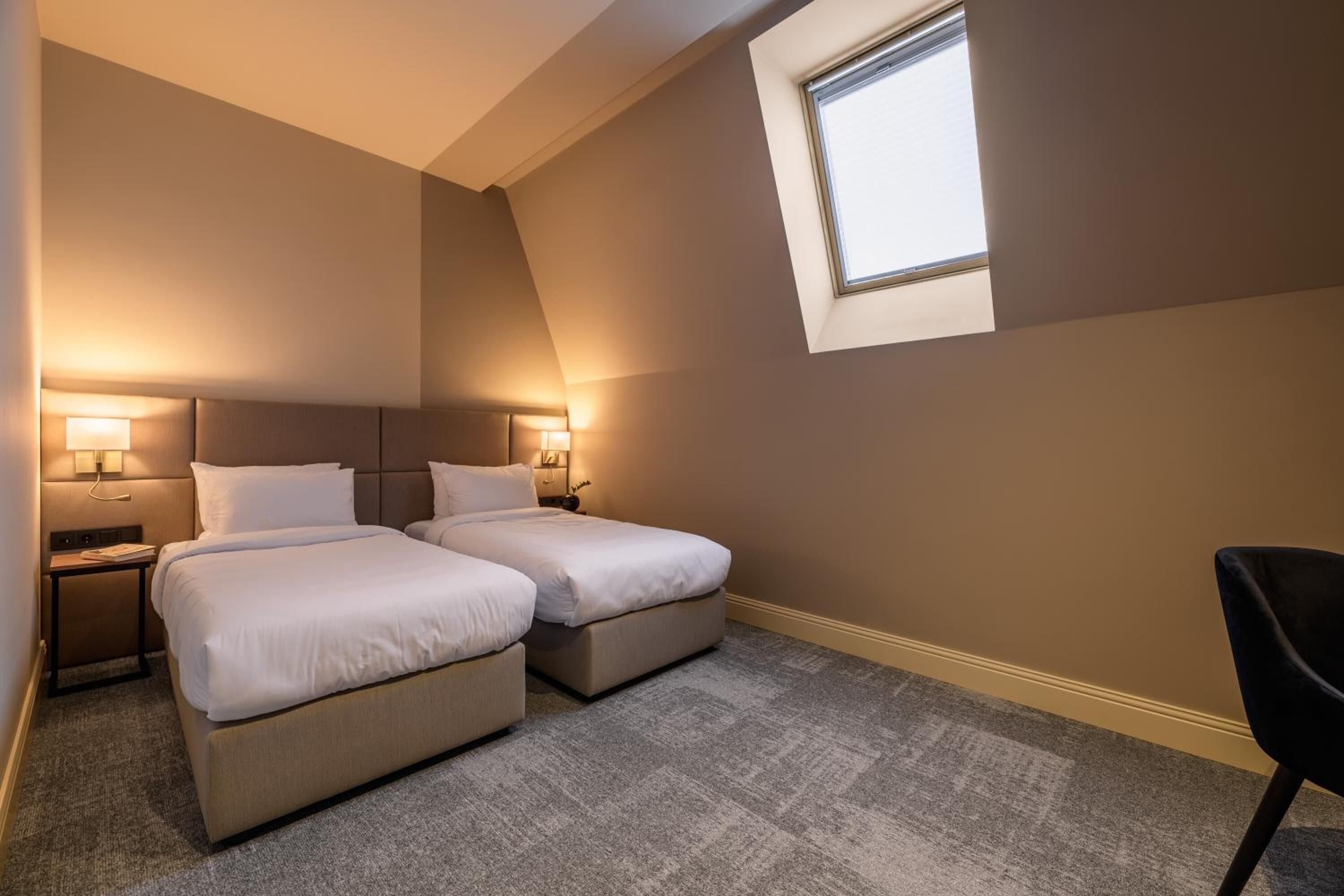 A hotel room with two beds, a small table, a chair, and a window.
