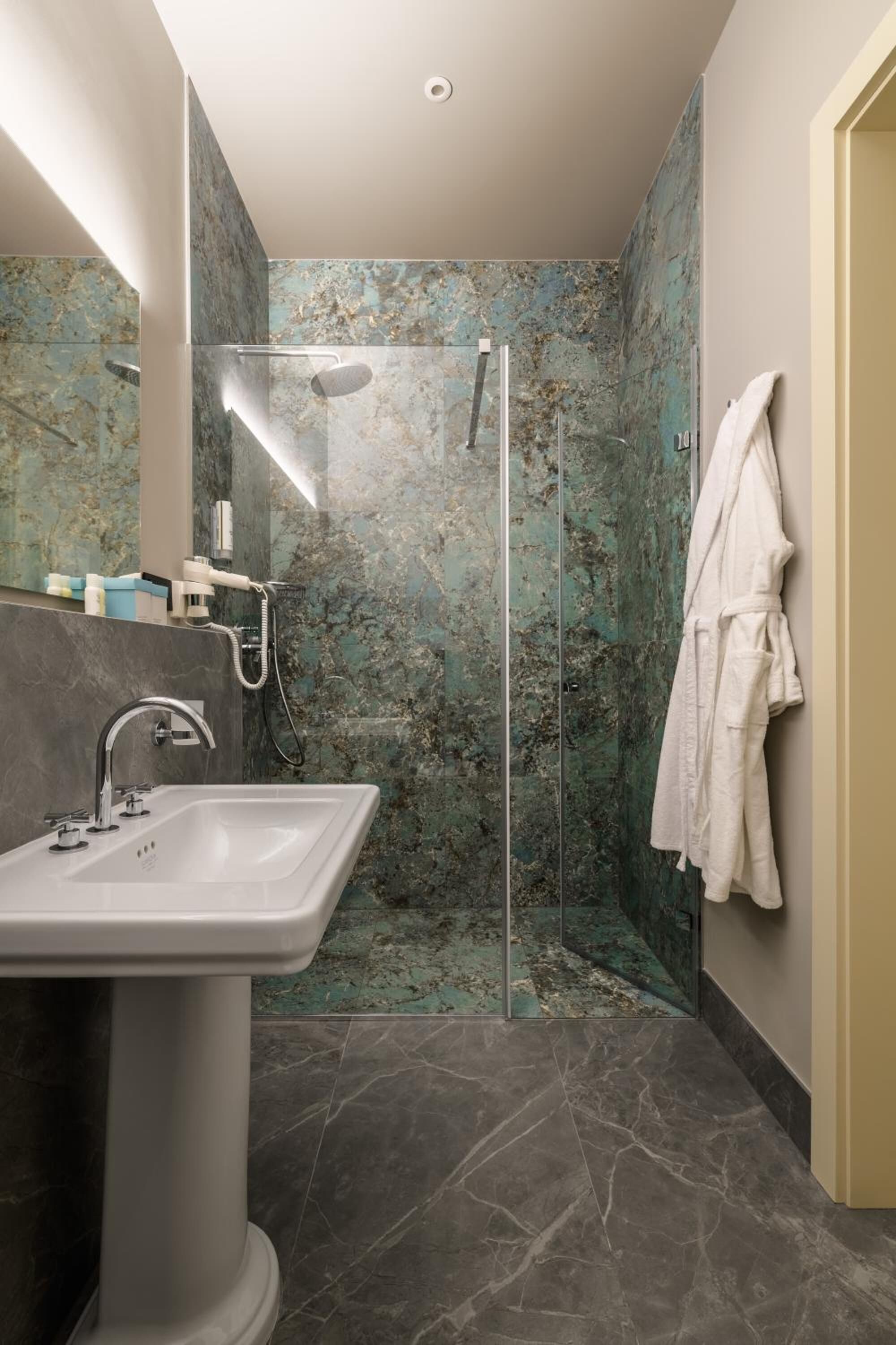 Un baño moderno con ducha de vidrio, lavamanos sobre pedestal y una bata blanca colgada en la pared.