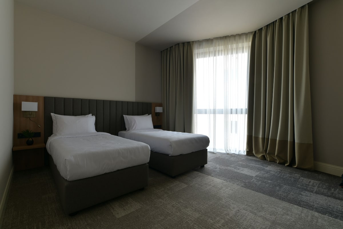 A hotel room with two single beds, a large window with curtains, a wooden headboard, and a small bedside table with a lamp.