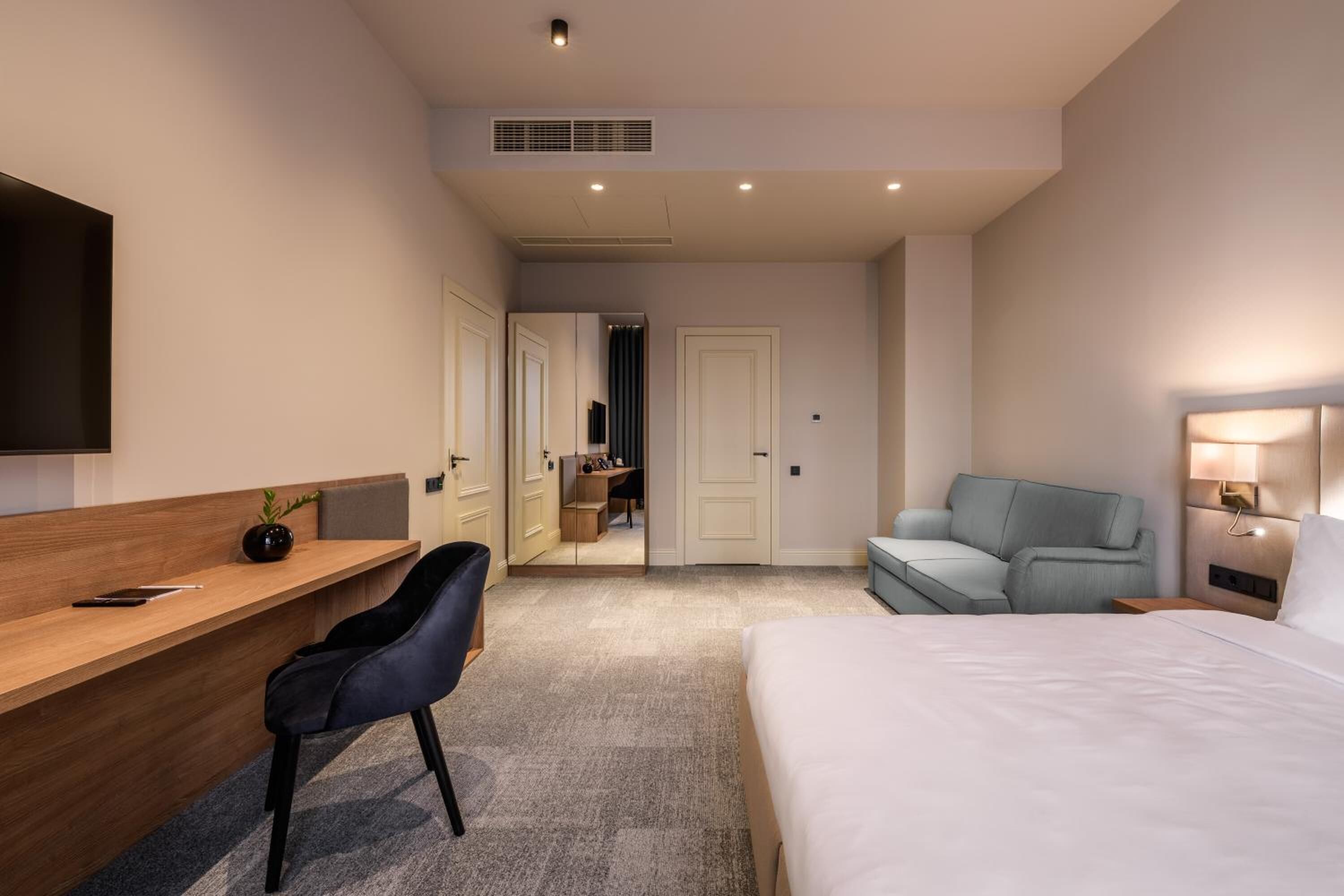 Quarto de hotel moderno com uma cama, uma mesa com cadeira, um sofá e uma TV fixada na parede.