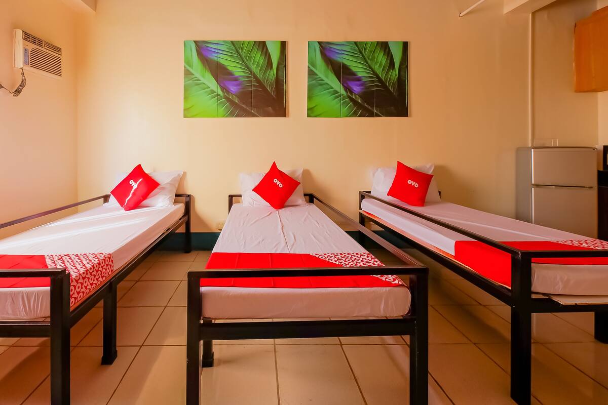 A room with three single beds, each with a red pillow and a white bedspread. There are two framed pictures on the wall above the beds.