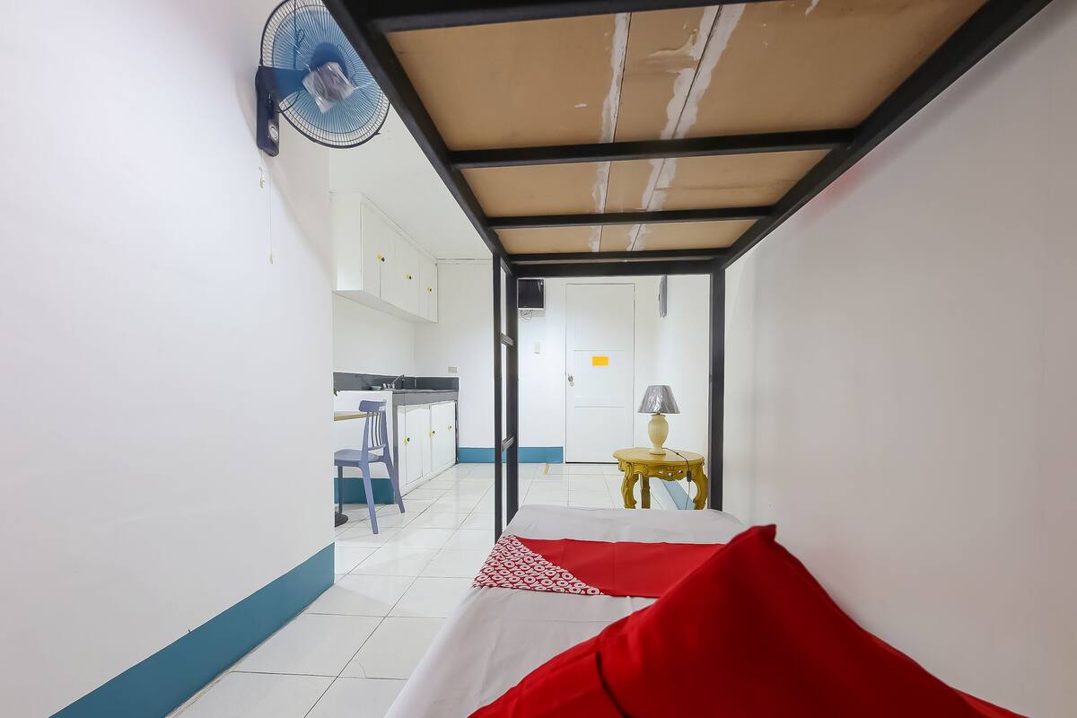 A narrow corridor leading to a room with a bed, a fan, a table with a lamp, and a kitchen area with a sink and cabinets.