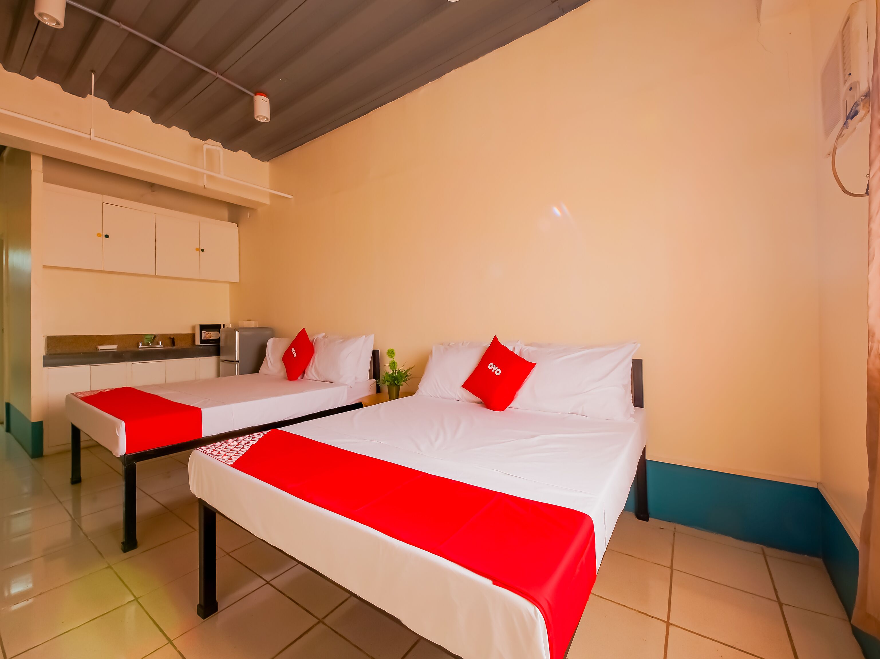 A hotel room with two beds, each with a red pillow, white bedding, and a red runner. There is a kitchenette with a fridge and a sink.