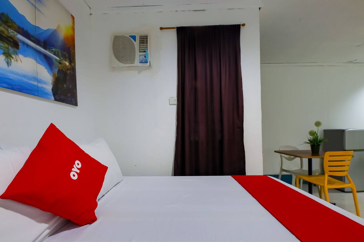 A hotel room with a bed, a red pillow, a painting on the wall, an air conditioning unit, a curtain, a desk with a chair, and a small plant.