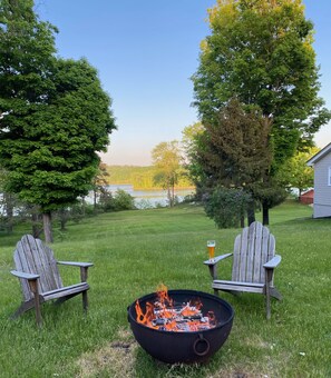 Ein Garten mit einem Feuerkorb, zwei Adirondack-Stühlen und einem Glas Bier auf einem kleinen Tisch.