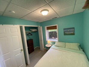 Ein Schlafzimmer mit türkisfarbenen Wänden, einer weißen Tür, einem Bett mit weißer Bettwäsche, einem Nachttisch mit einer Lampe, einem Fenster mit Blick auf Grünflächen und einem Bild an der Wand.