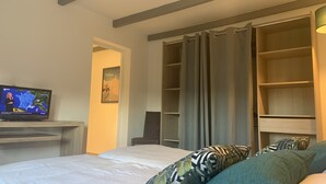 Quarto de hotel com cama, televisão exibindo um noticiário, guarda-roupa e uma janela grande com cortinas.