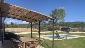 Um pátio externo com mesa e cadeiras de madeira, uma área de estar coberta e uma piscina.
