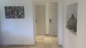 Um corredor com piso de azulejos, duas portas brancas, uma pintura de azeitonas e um vaso na parede.