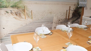 A dining table set with white plates, glasses, and cutlery, overlooking a beach with a wooden deck and a view of the ocean.